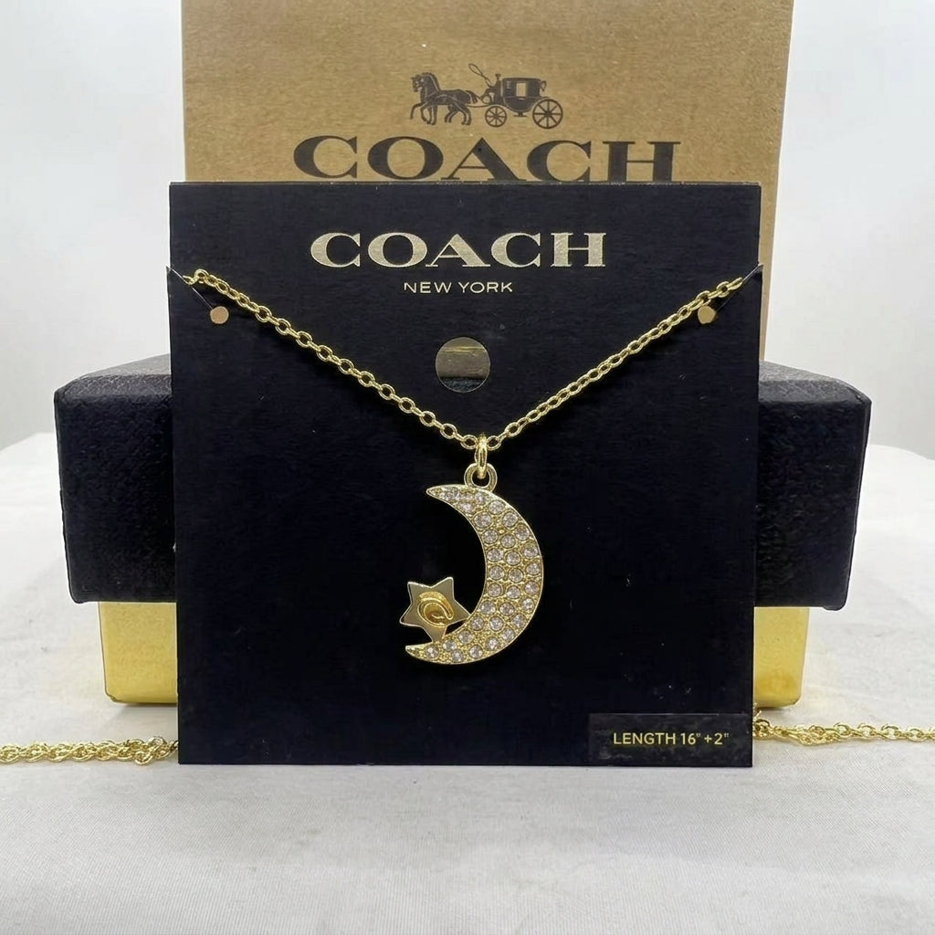 A gold-tone Coach necklace with a crescent moon and star pendant covered in small clear stones, displayed on a black Coach New York card with a kraft Coach box behind it.