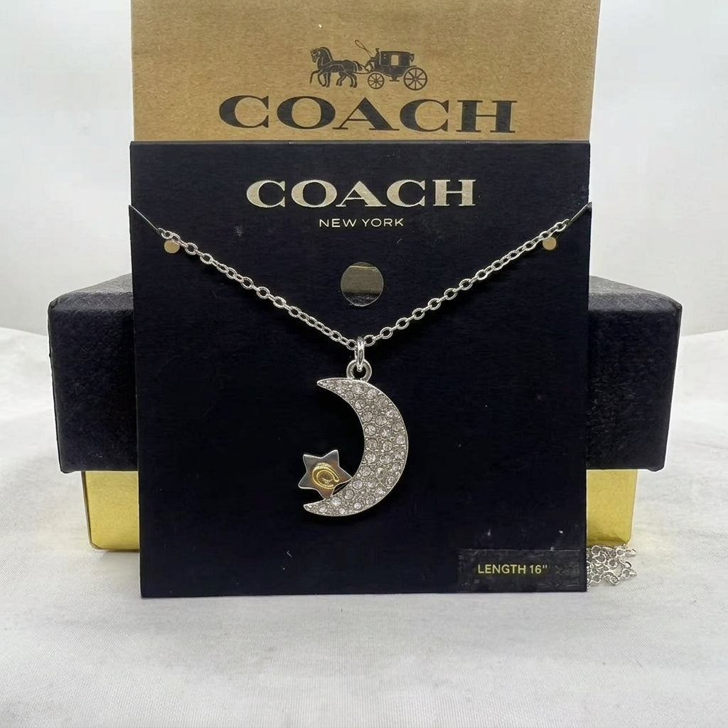A silver-tone Coach necklace with a crescent moon and star pendant, displayed on a black Coach New York card with a gold Coach box behind it.