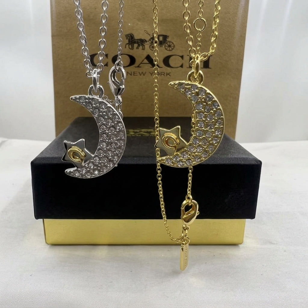 Two Coach New York crescent moon and star necklaces displayed in front of their branded packaging. One necklace is silver with clear rhinestones and a gold star center, and the other is gold with clear rhinestones and a gold star center.