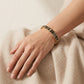 A gold bangle bracelet with black enamel and the word 'COACH' repeated in gold lettering, worn on a woman's wrist resting on a beige textured surface.