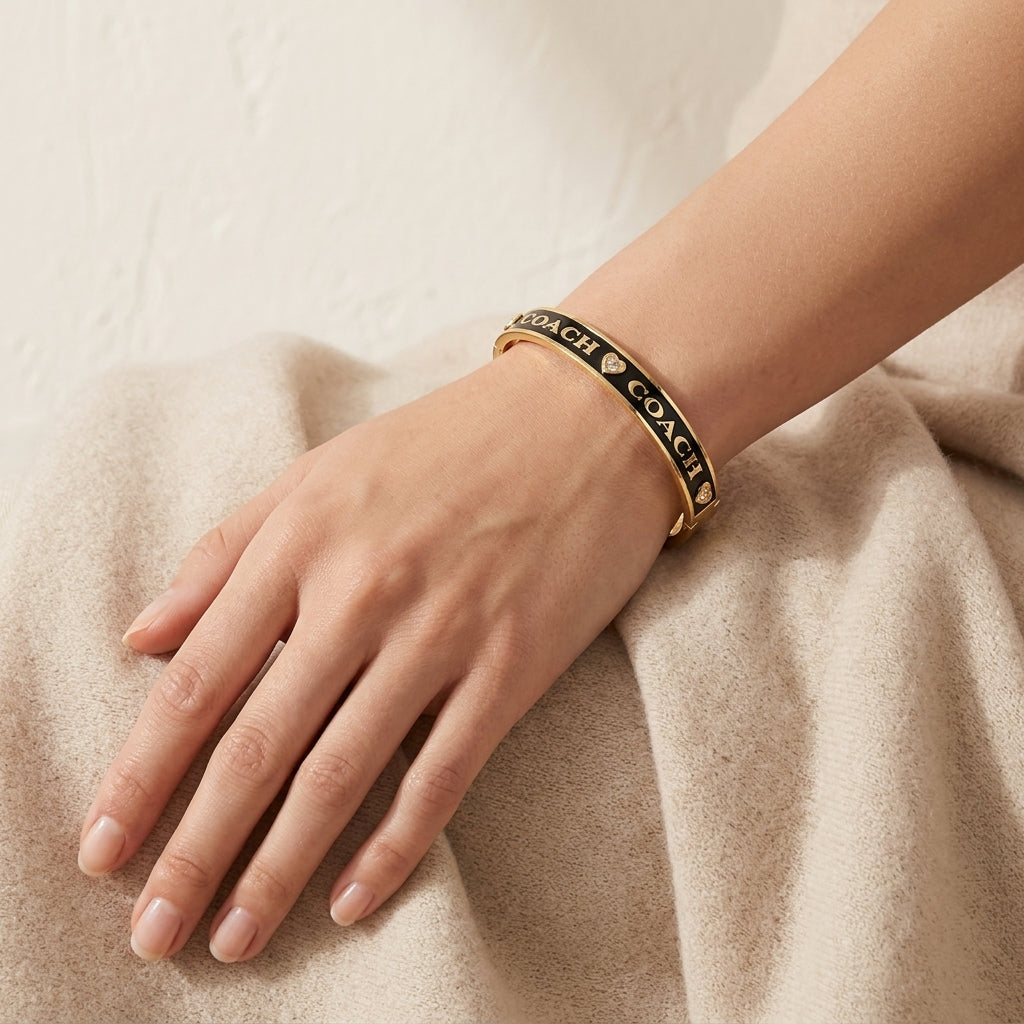 A gold bangle bracelet with black enamel and the word 'COACH' repeated in gold lettering, worn on a woman's wrist resting on a beige textured surface.