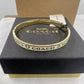A gold-tone Coach bangle bracelet with white enamel and black lettering, displayed on top of its black textured brand box.