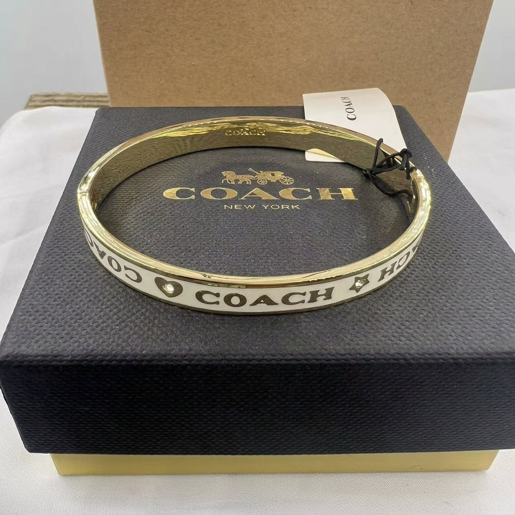 A gold-tone Coach bangle bracelet with white enamel and black lettering, displayed on top of its black textured brand box.