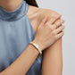 A close-up lifestyle shot of a model wearing a gold bangle bracelet and a gold ring on her wrist, wearing a blue satin sleeveless top against a light gray background.