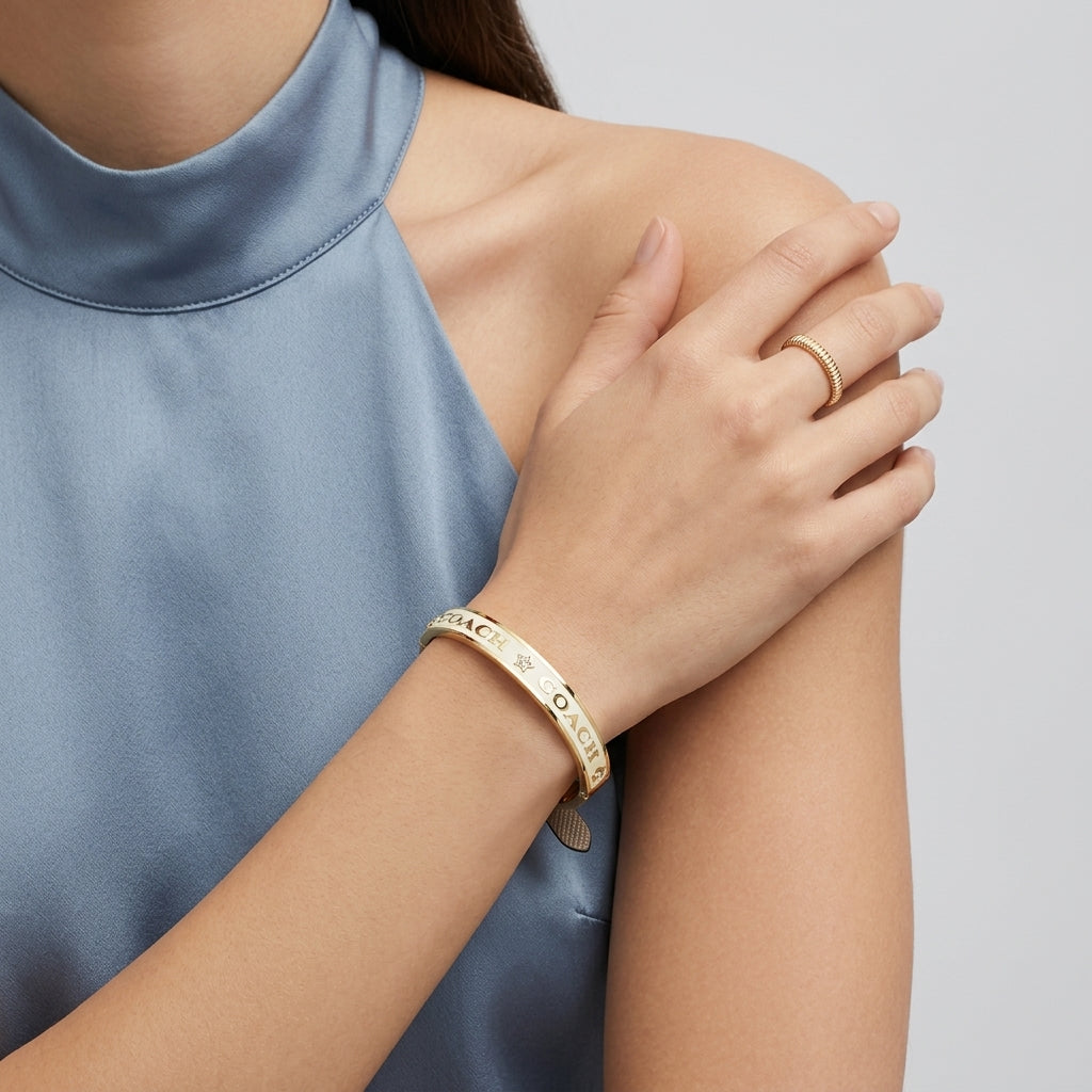 A close-up lifestyle shot of a model wearing a gold bangle bracelet and a gold ring on her wrist, wearing a blue satin sleeveless top against a light gray background.