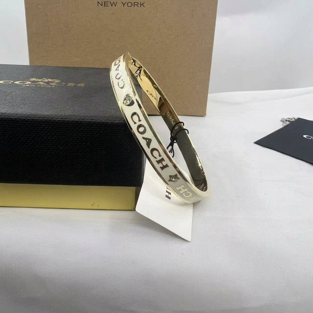 A gold bangle bracelet with black Coach logo lettering and diamond motifs, displayed in front of its black and gold presentation box on a white surface.