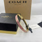 A gold-tone Coach bangle bracelet with pink enamel and gold lettering, displayed next to its black and gold Coach box and key on a white surface.
