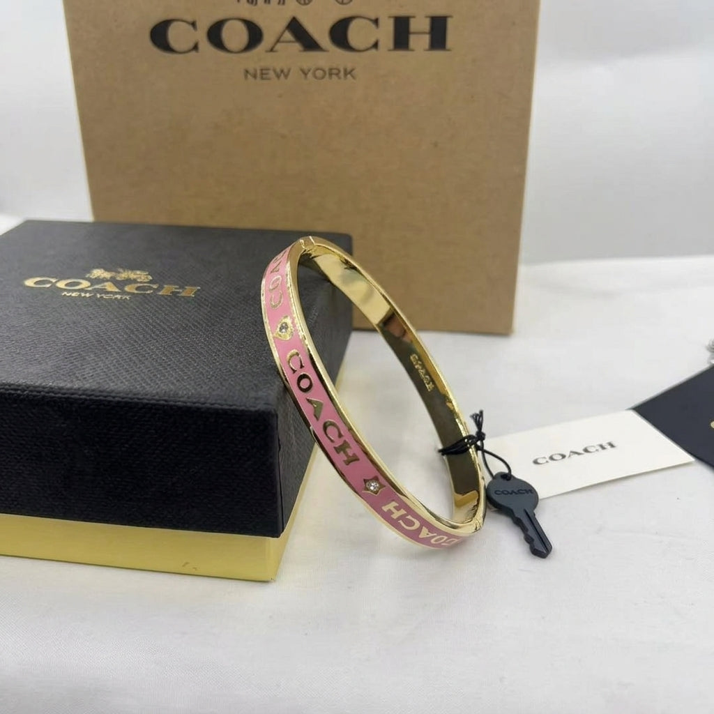 A gold-tone Coach bangle bracelet with pink enamel and gold lettering, displayed next to its black and gold Coach box and key on a white surface.