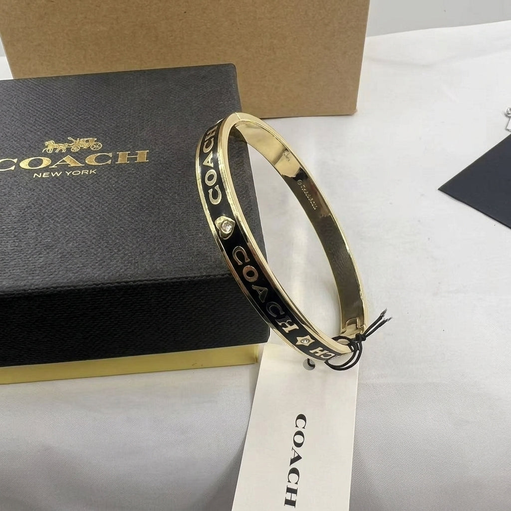 A gold-tone Coach bangle bracelet with black lettering and small crystal accents, displayed next to a black Coach New York box and branded hangtag on a white surface.