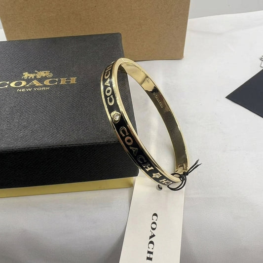A gold-tone Coach bangle bracelet with black lettering and small crystal accents, displayed next to a black Coach New York box and branded hangtag on a white surface.