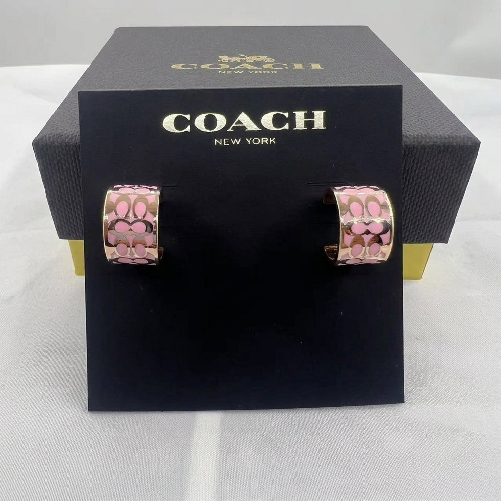 A pair of Coach New York hoop earrings with a pink and gold floral pattern, displayed on a black branded card in front of a black Coach box.