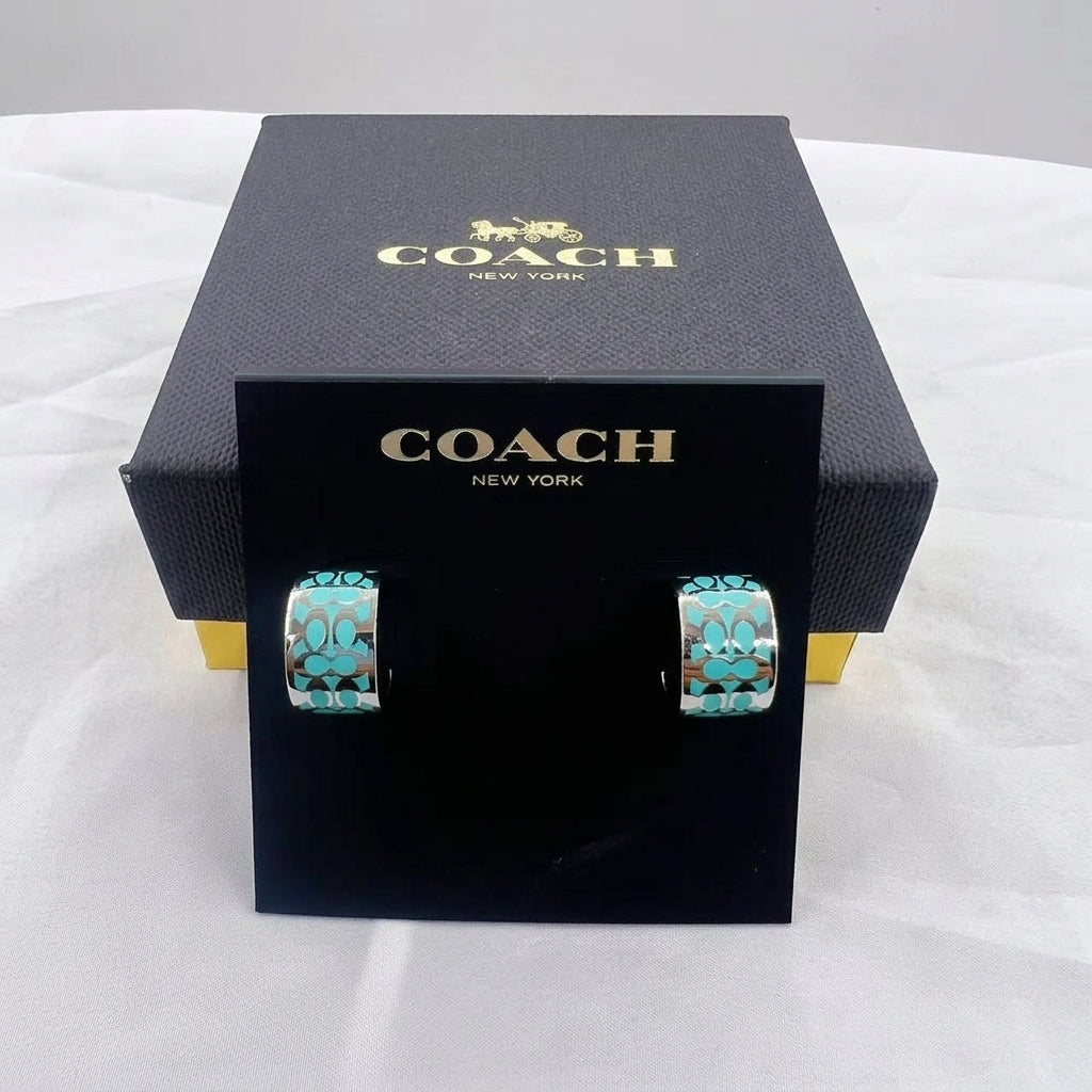 Coach New York hoop earrings with turquoise and gold patterned inlay, displayed on a black branded card in front of a black Coach box on a white fabric background.