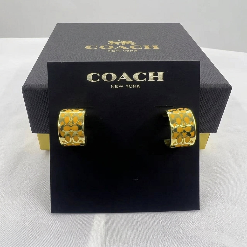 A pair of gold hoop earrings with an embossed Coach logo pattern, displayed on a black Coach New York card stand in front of their black presentation box.