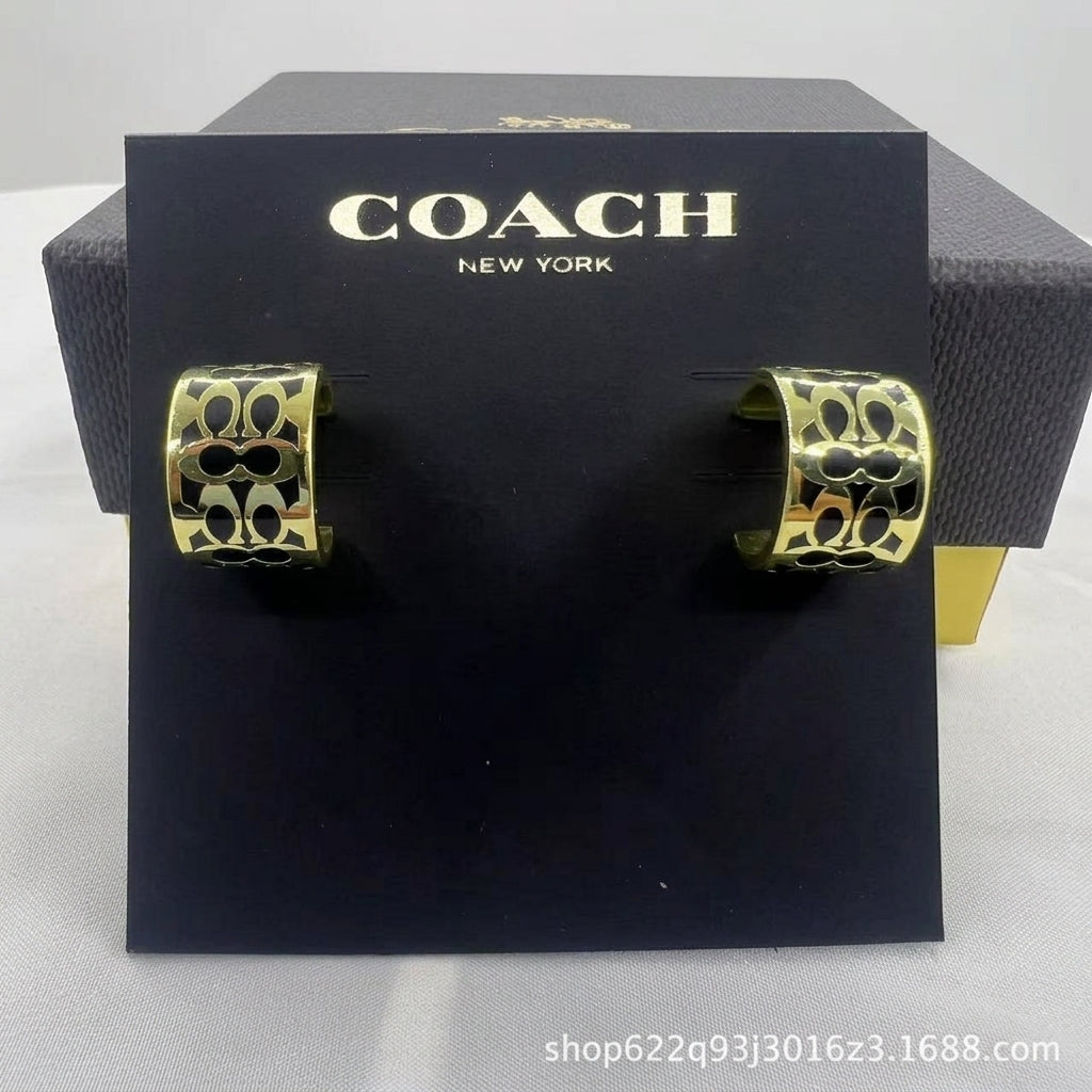 A pair of gold-tone hoop earrings with black interlocking monogram cutouts, displayed on a black Coach New York branded card, with the Coach box behind it.