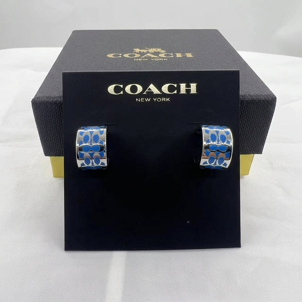 A pair of Coach huggie hoop earrings with blue and silver floral enameled details, displayed on a black Coach New York branded card stand in front of their black presentation box.