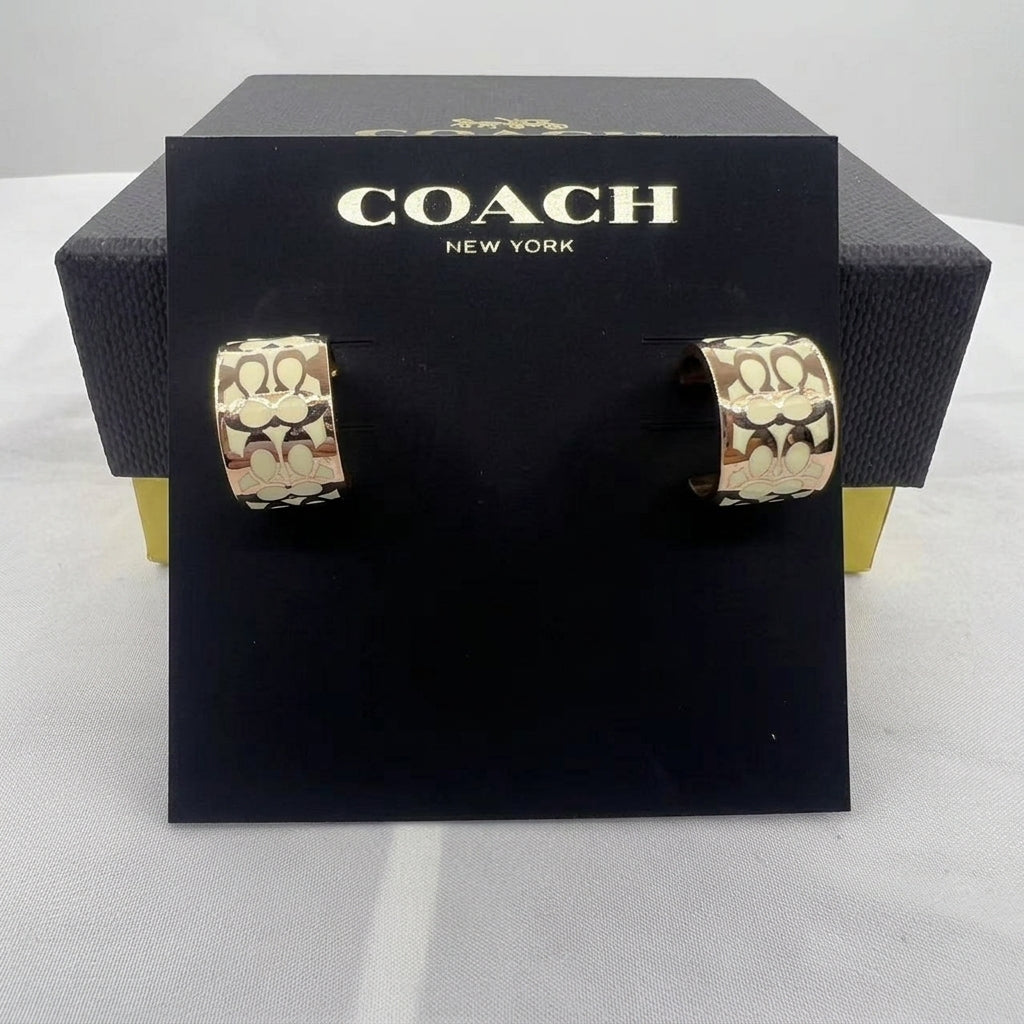 A pair of Coach New York hoop earrings with a gold-tone and cream signature 'C' pattern, displayed on a black branded card stand in front of a black Coach box.
