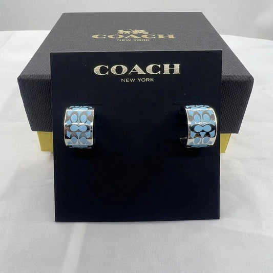 A pair of Coach New York hoop earrings with a light blue and silver signature pattern, displayed on a black branded card stand in front of a black Coach box.