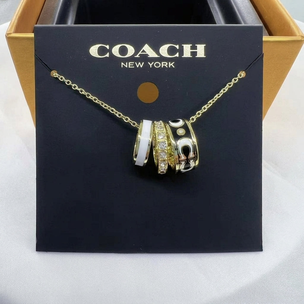 A Coach New York gold chain necklace with three interlocking cylindrical charms, displayed on a black branded card insert. The charms are gold-tone with white, black, and clear crystal details, set against a gold and black Coach box.