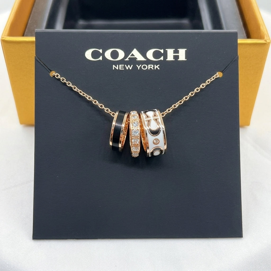 A Coach New York necklace displayed on a black branded card, with three interlocking ring-shaped charms in rose gold, black, and white with crystal detailing, set against a gold box background.