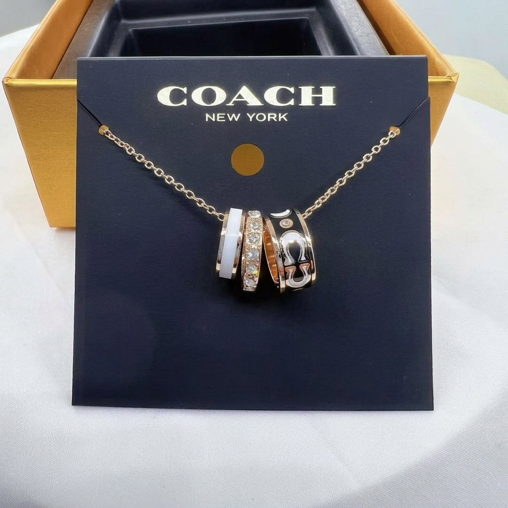 A Coach New York necklace with three interlocking charm rings in rose gold, white, and black, displayed on a black branded card in front of a gold box.