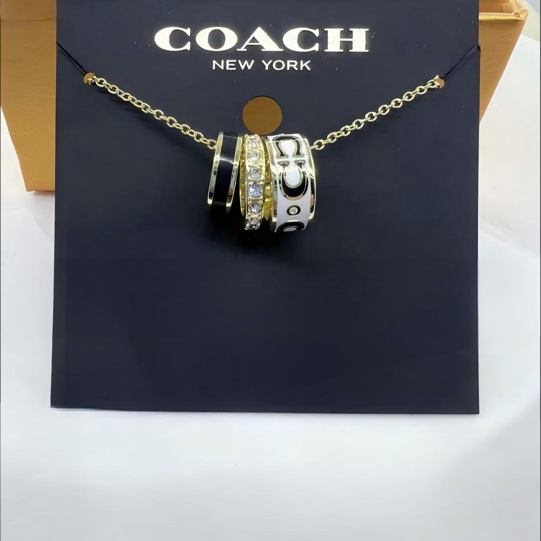 A Coach New York necklace with a gold chain and a multi-spoke charm featuring black, white, and clear crystal details, displayed on a black branded card.