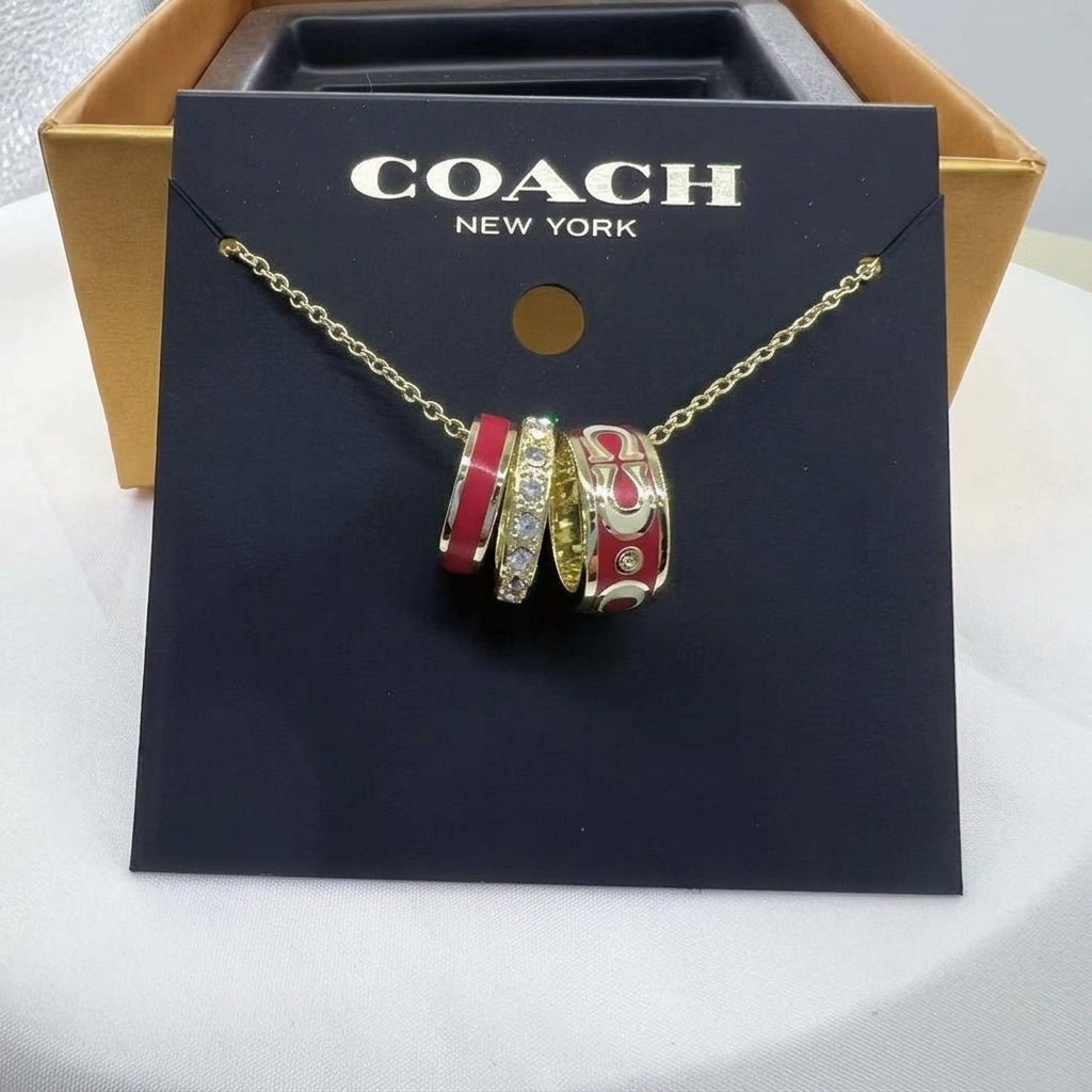 A Coach New York necklace with a gold chain and a multi-strand charm pendant featuring red, white, and gold sections, displayed on a black branded card insert in front of a gold box.