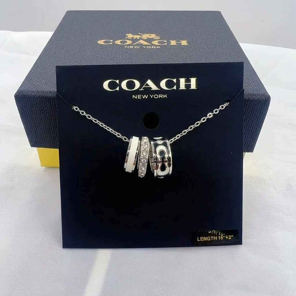 A Coach New York silver charm necklace displayed on a black branded card, with the dark Coach box behind it on a white fabric surface.