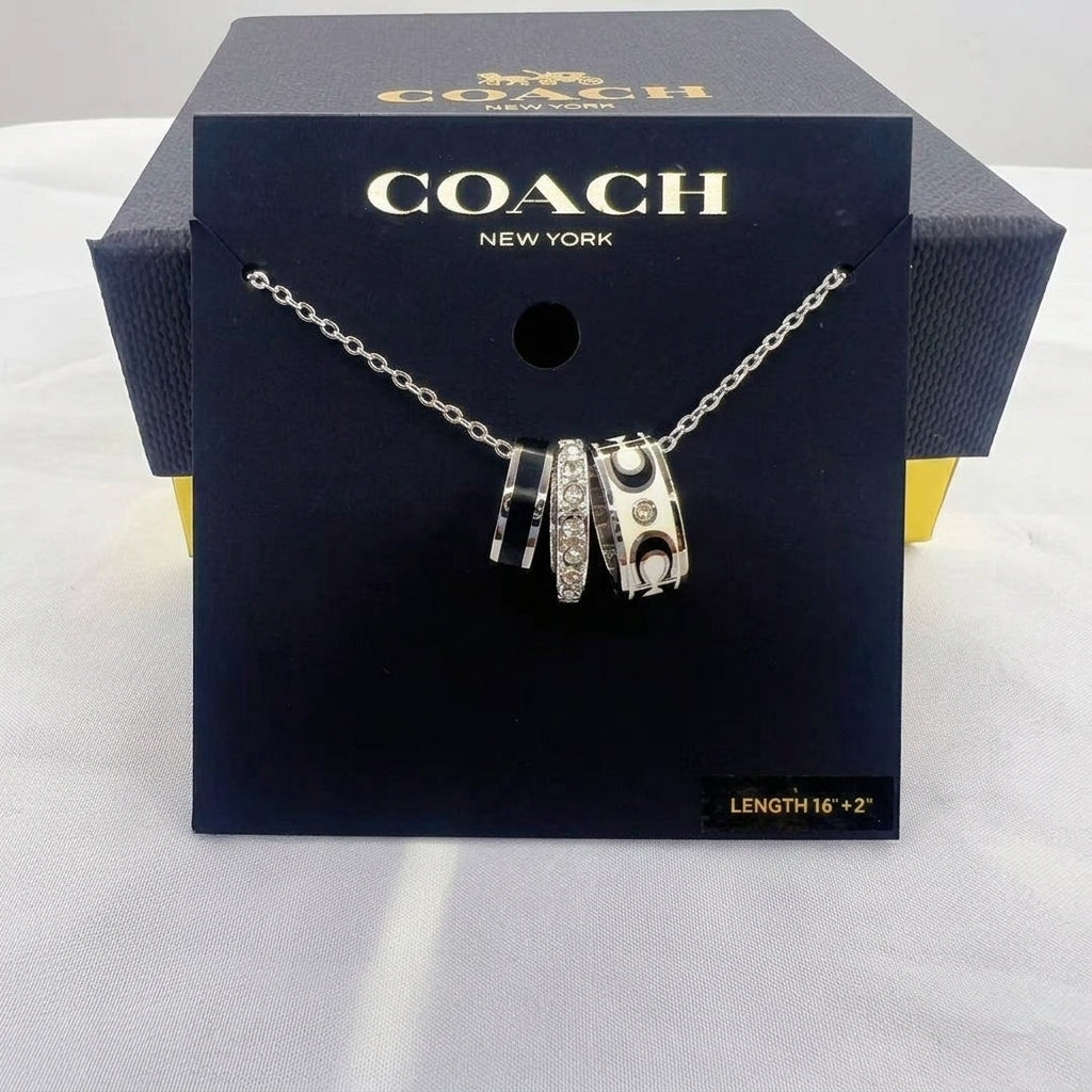 A Coach New York necklace displayed on a branded black box. The necklace has three silver-toned cylindrical charms with black enamel sections, one set with clear stones, and one with a gold-tone 'C' logo, hanging from a silver chain.