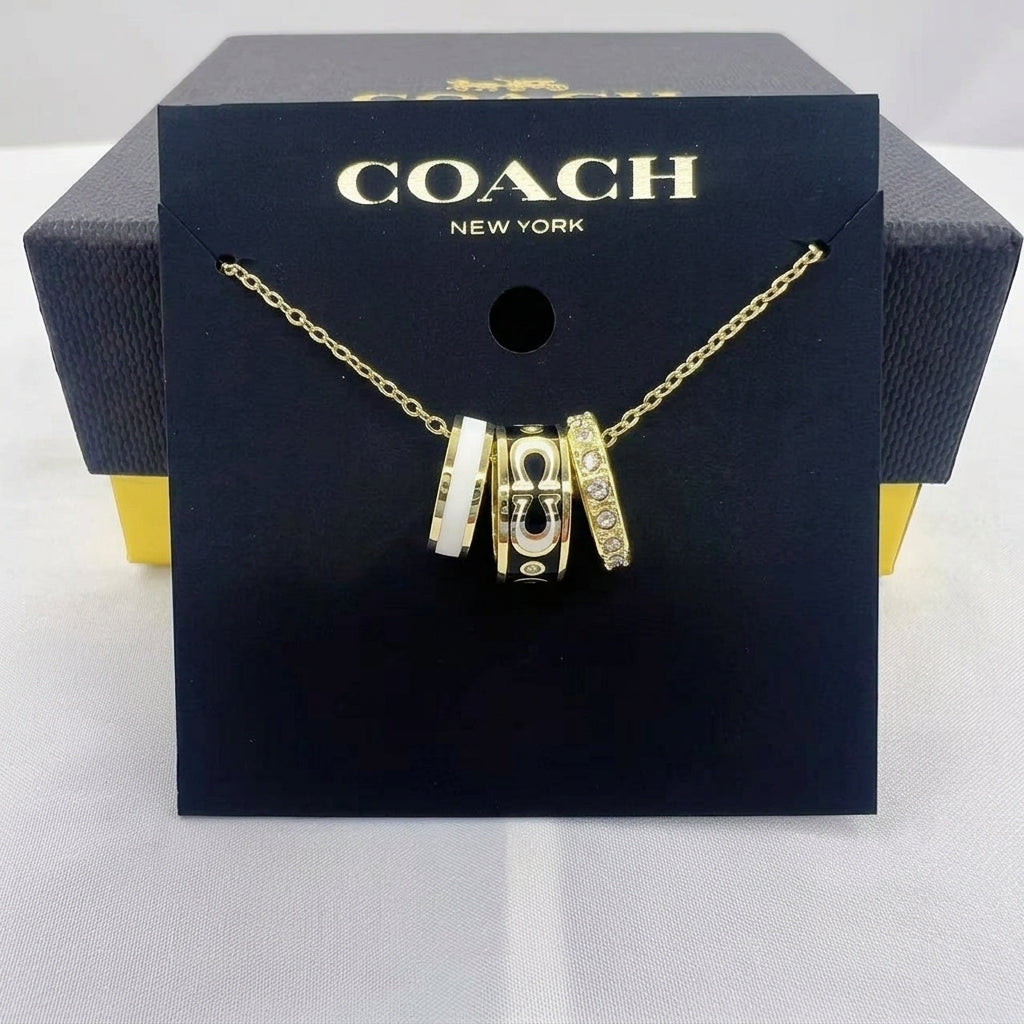 A gold-tone Coach necklace with three interlocking padlock charms, displayed on a black Coach New York branded card and box.