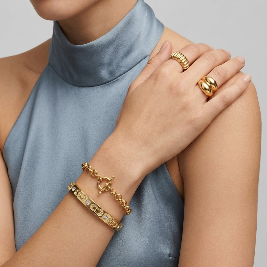 A close-up of a model wearing gold jewelry, including a textured gold ring, a polished gold signet-style ring, and two gold bracelets on one wrist, paired with a light blue satin high-neck top against a neutral gray background.