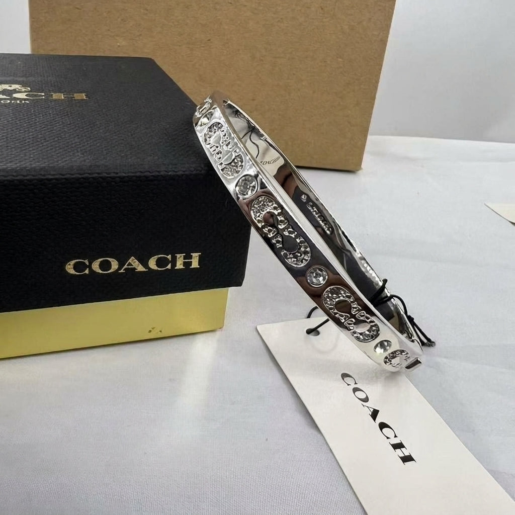 A silver-tone Coach bangle bracelet with circular crystal-embellished motifs, displayed next to its black and gold branded box and a white COACH tag on a light surface.