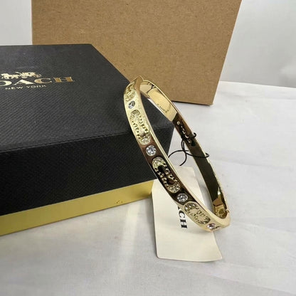 A gold-tone Coach bangle bracelet with clear rhinestone accents, displayed next to its black and gold Coach box and hangtag on a white surface.