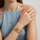A close-up of a model wearing gold jewelry, featuring a thick gold ring on one finger and a stack of three gold bracelets on the wrist, set against a light blue sleeveless top and a neutral gray background.