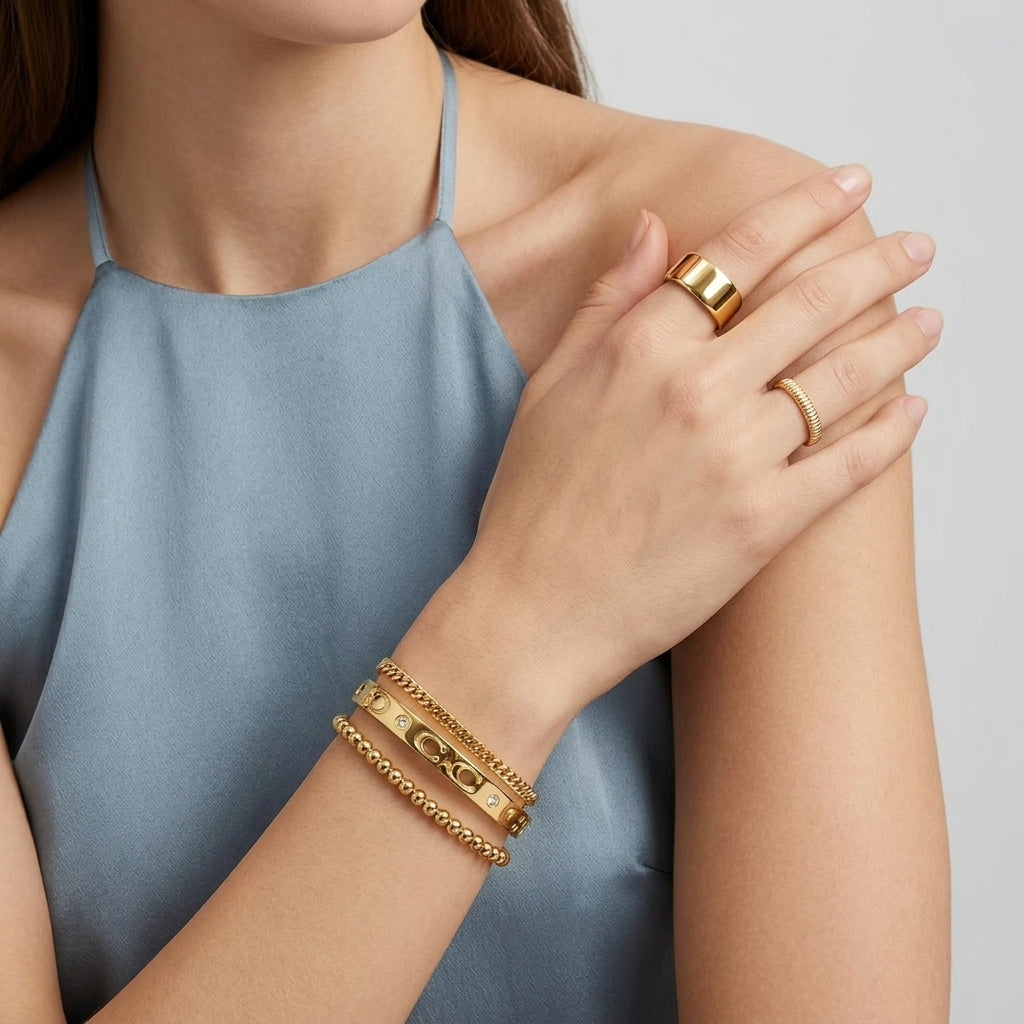 A close-up of a model wearing gold jewelry, featuring a thick gold ring on one finger and a stack of three gold bracelets on the wrist, set against a light blue sleeveless top and a neutral gray background.