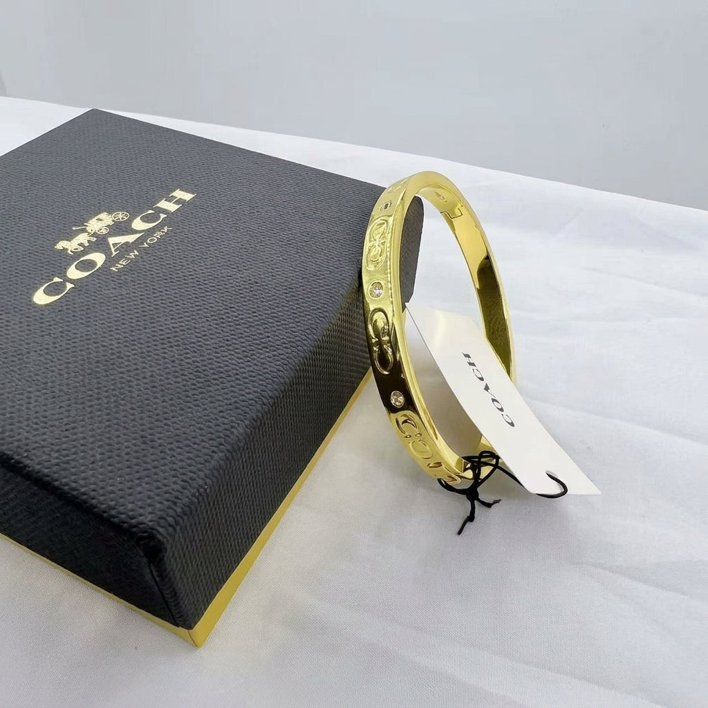 A gold-tone Coach bangle bracelet with engraved details and small clear stones, shown next to its black and gold Coach New York gift box on a white fabric surface.