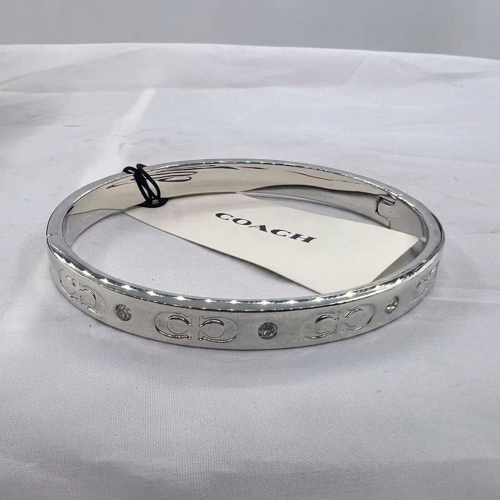 A silver-tone Coach bangle bracelet with engraved 'CO' logos and small crystal accents, shown with a white Coach paper tag on a white fabric background.