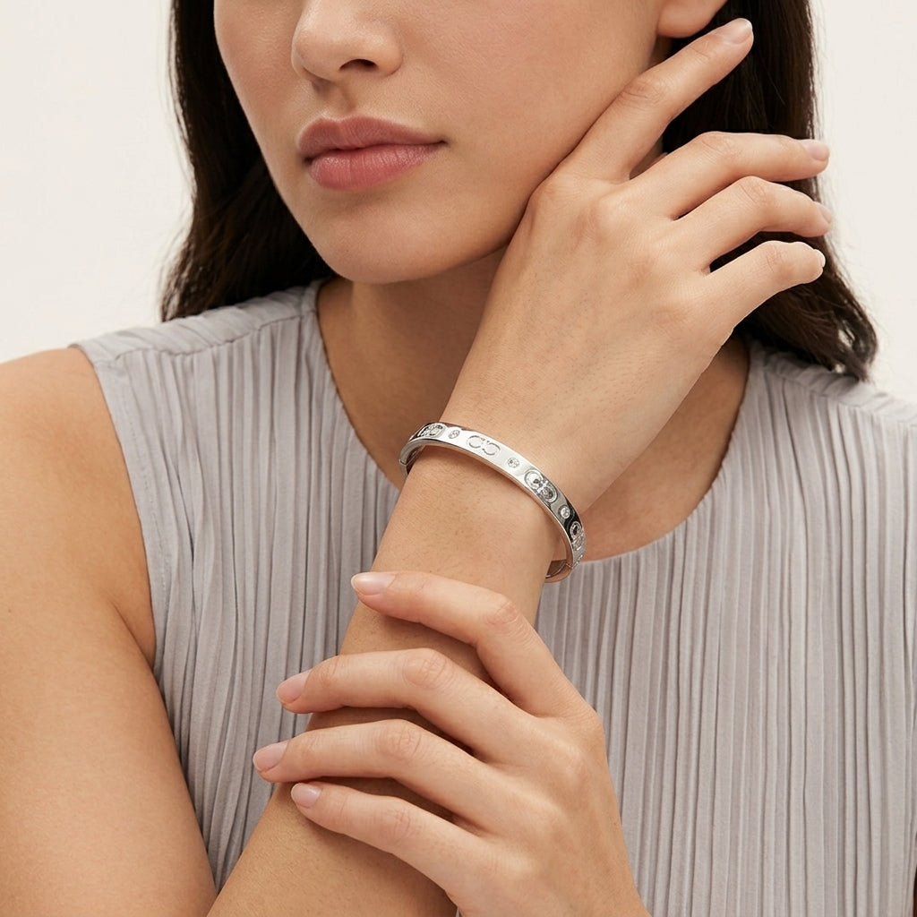 A close-up of a woman wearing a polished silver bangle bracelet with engraved lettering, paired with a light gray pleated top.