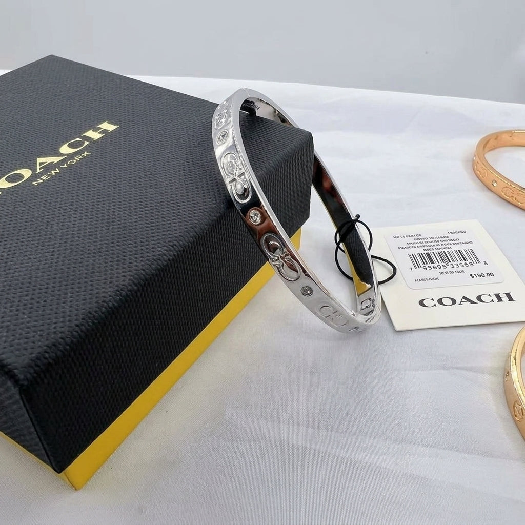 A silver-tone Coach bangle bracelet with engraved logos and small clear stones, displayed next to its black and yellow Coach box and a price tag on a white fabric surface.