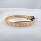 A gold-tone Coach bangle bracelet with its original white paper tag attached, displayed on a white fabric surface.