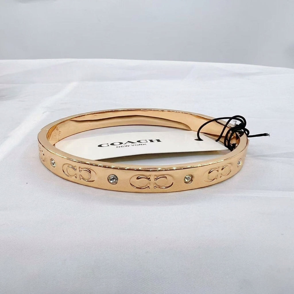 A gold-tone Coach bangle bracelet with its original white paper tag attached, displayed on a white fabric surface.