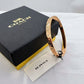 A rose gold bangle bracelet with a floral pattern, shown next to its black and gold Coach New York box and a branded tag on a white surface.
