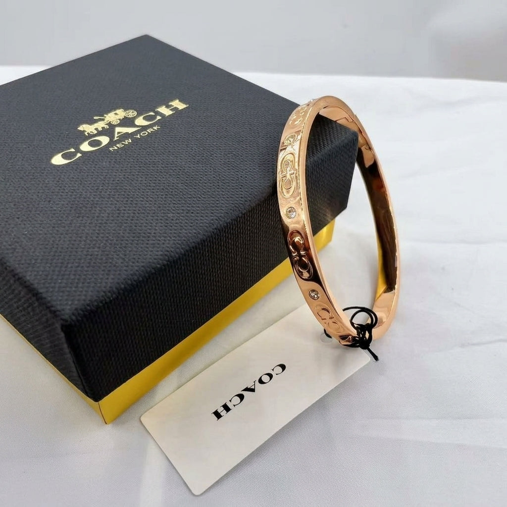 A rose gold bangle bracelet with a floral pattern, shown next to its black and gold Coach New York box and a branded tag on a white surface.