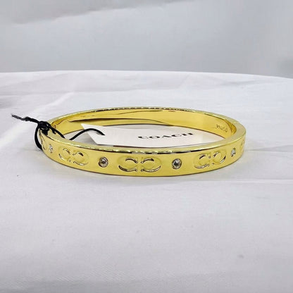 A gold bangle bracelet with engraved interlocking 'C' logos and small clear rhinestone accents, shown on a white fabric surface with a Coach tag attached.
