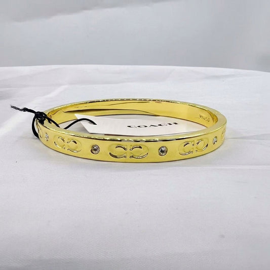 A gold bangle bracelet with engraved interlocking 'C' logos and small clear rhinestone accents, shown on a white fabric surface with a Coach tag attached.