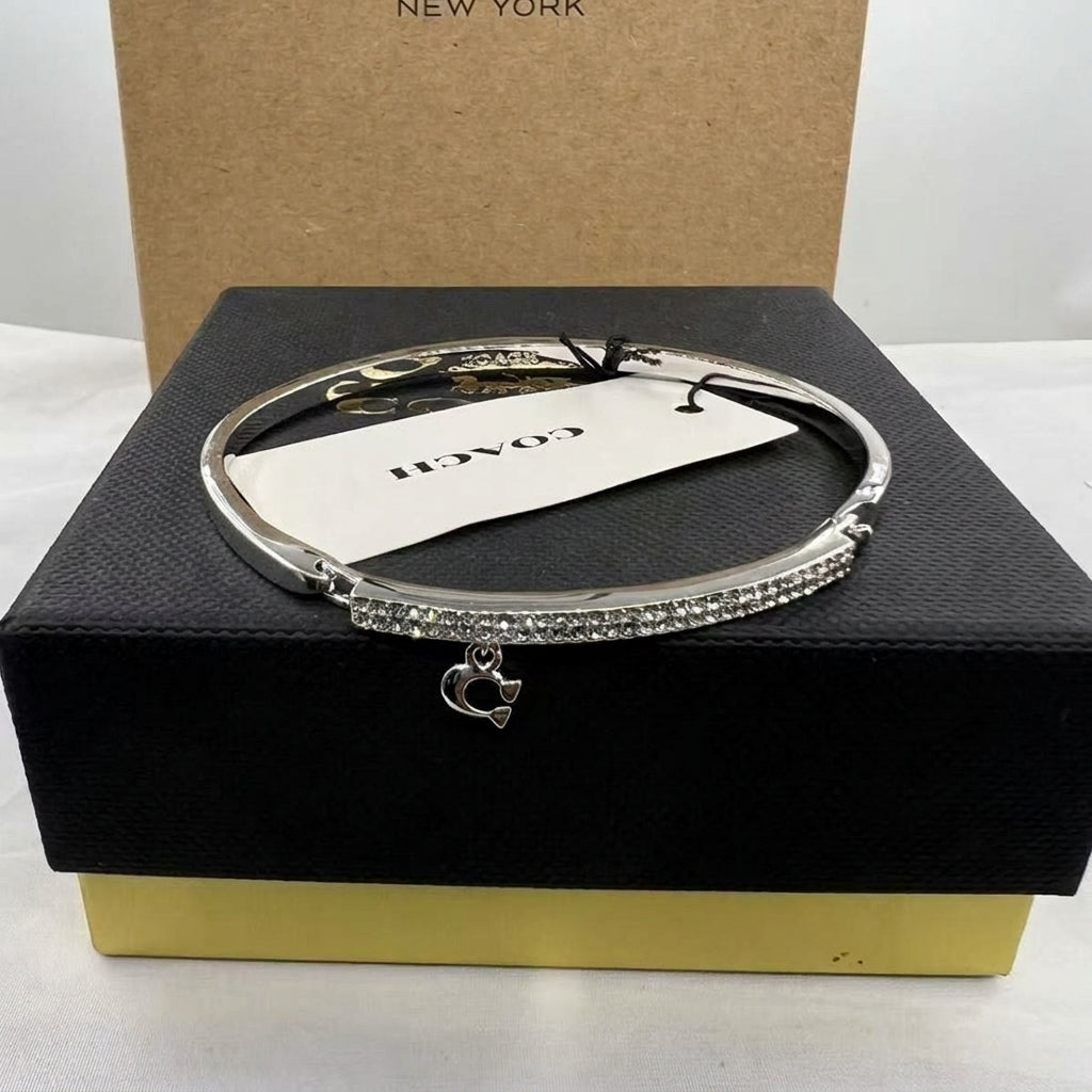 A silver-tone Coach bangle bracelet with a crystal-encrusted band and a small dangling Coach logo charm, displayed on a black Coach presentation box with a gold base.