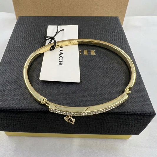 A gold-tone Coach bangle bracelet with a crystal-encrusted bar and small logo charm, displayed on a black Coach box with its white hangtag.