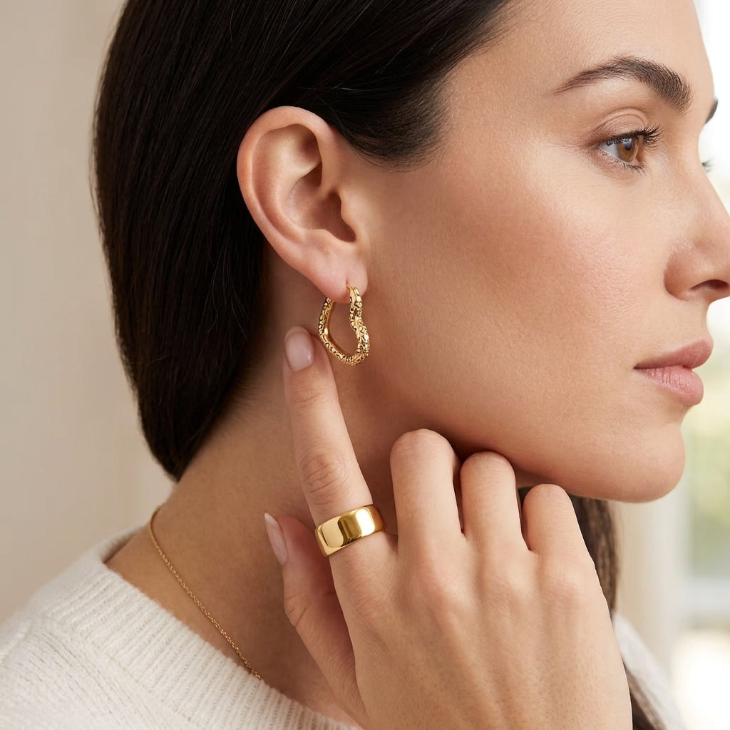 A close-up side profile of a woman wearing a textured gold hoop earring and a matching gold ring, with a delicate gold necklace also visible against a soft neutral background.