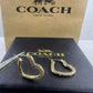 A pair of gold-tone Coach huggie hoop earrings with textured scrollwork, displayed on a black Coach box with tweezers, in front of a brown Coach New York box.