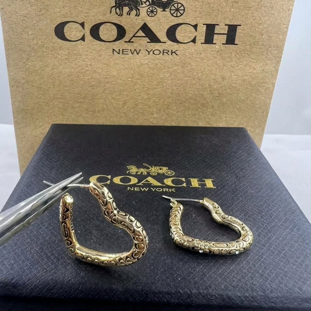 A pair of gold-tone Coach huggie hoop earrings with textured scrollwork, displayed on a black Coach box with tweezers, in front of a brown Coach New York box.