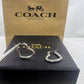 A pair of silver-tone Coach hoop earrings with pavé-set clear stones, shown with metal tweezers on top of a black Coach New York box, with a larger brown Coach New York box behind it.